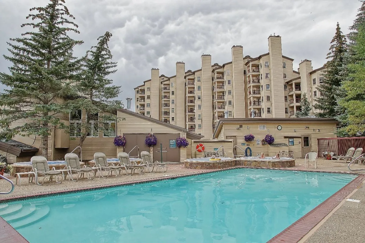 The image depicts the exterior of the property with a swimming pool surrounded by lounge chairs and decorative flowers. The building facade is visible in the background, blending seamlessly with the natural surroundings. The hot tub area is also featured, evoking a sense of relaxation and leisure after a day on the slopes.