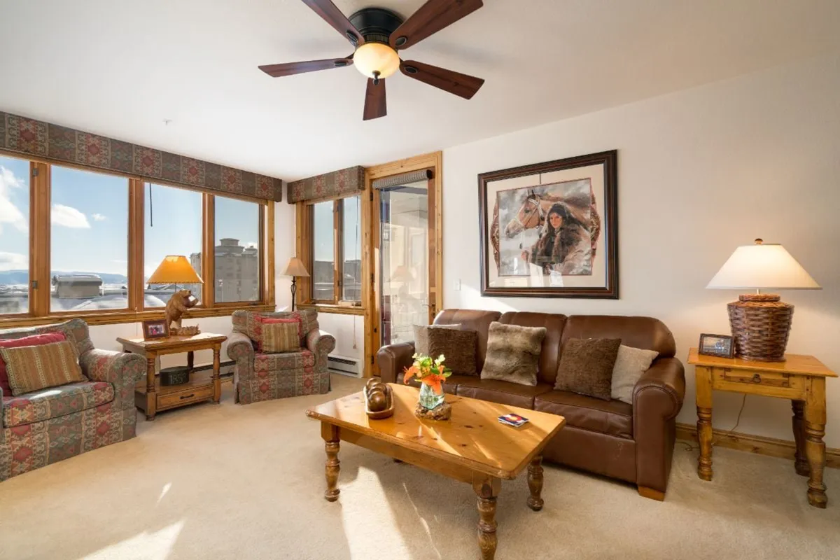 A cozy living space with large windows framing mountain views, a leather sofa, a wooden coffee table, and decorative artwork on the walls. The room conveys a welcoming transition from the outdoors, hinting at the proximity to mountain adventures.