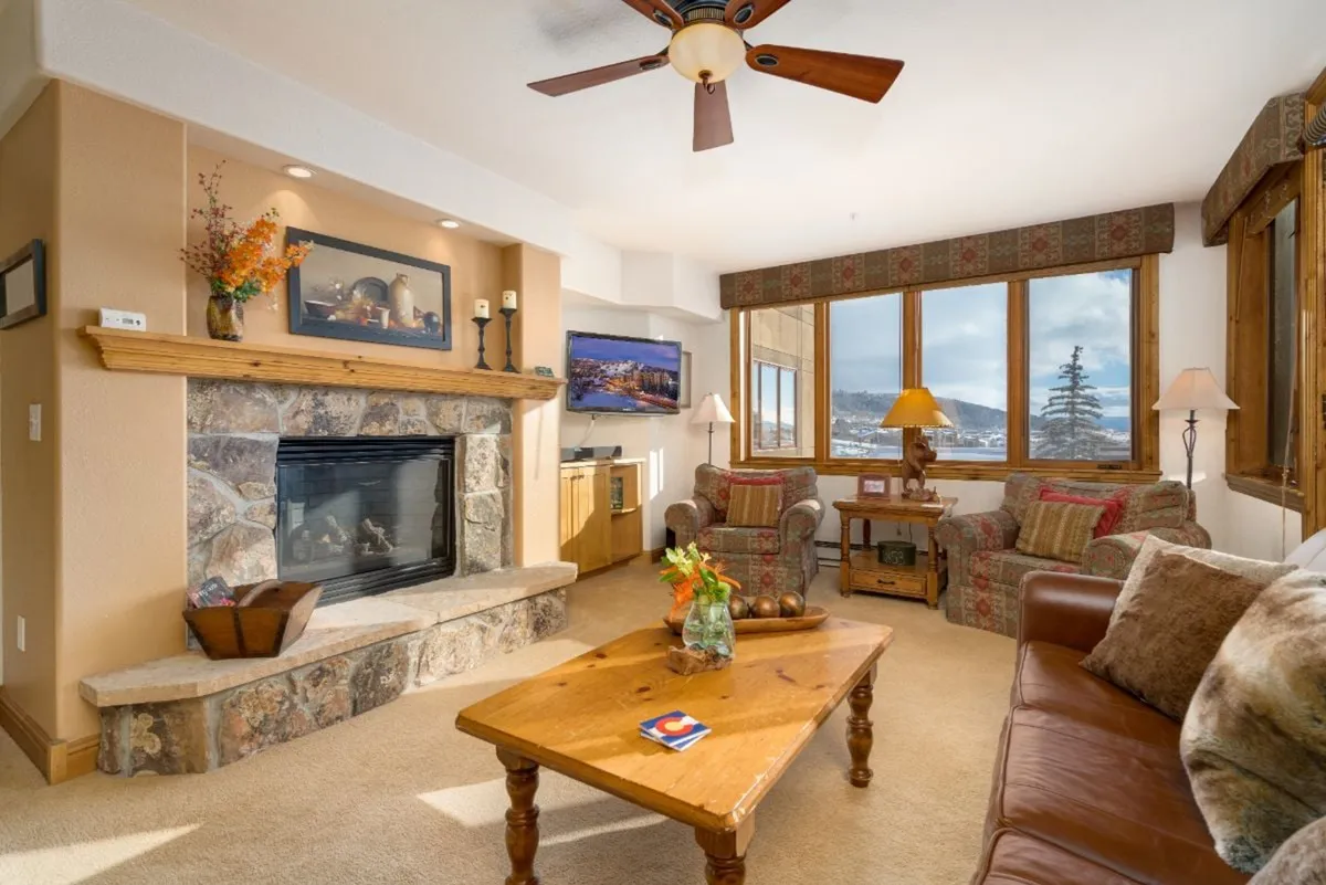 A welcoming living area featuring a plush couch and armchairs arranged around a cozy fireplace, with large windows allowing sunlight to pour in. Decorative touches and a coffee table add warmth, while the spaciousness supports socializing or quiet relaxation.