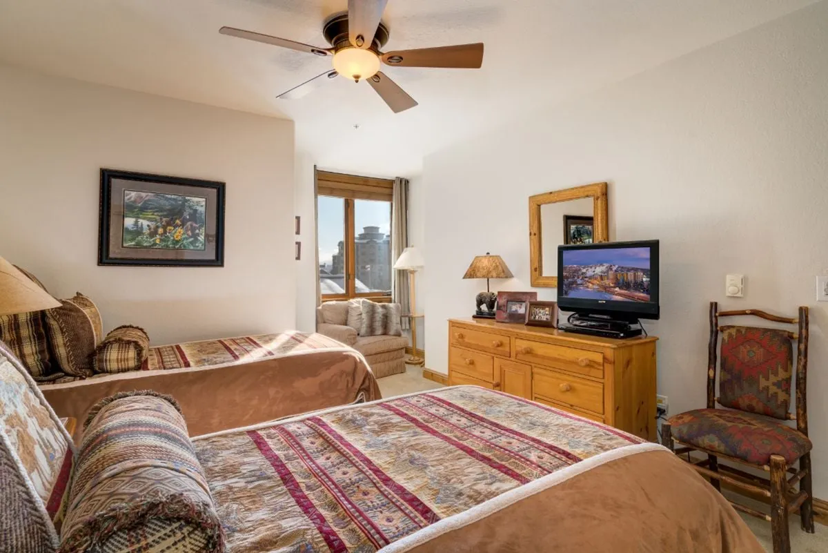 A bright and comfortable bedroom with two twin beds featuring decorative bedspreads, a wooden dresser with a TV, a sunlit window, and tasteful artwork. The space feels flexible and functional, ideal for families or friends traveling together.