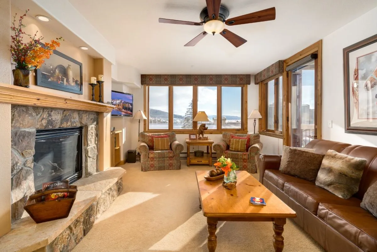 This inviting condo living room features a glowing fireplace, plush seating arranged for conversation, expansive windows that frame mountain sunsets, and a wooden coffee table adorned with fresh flowers. The space exudes warmth and comfort, perfect for après-ski relaxation or memorable evenings with loved ones.