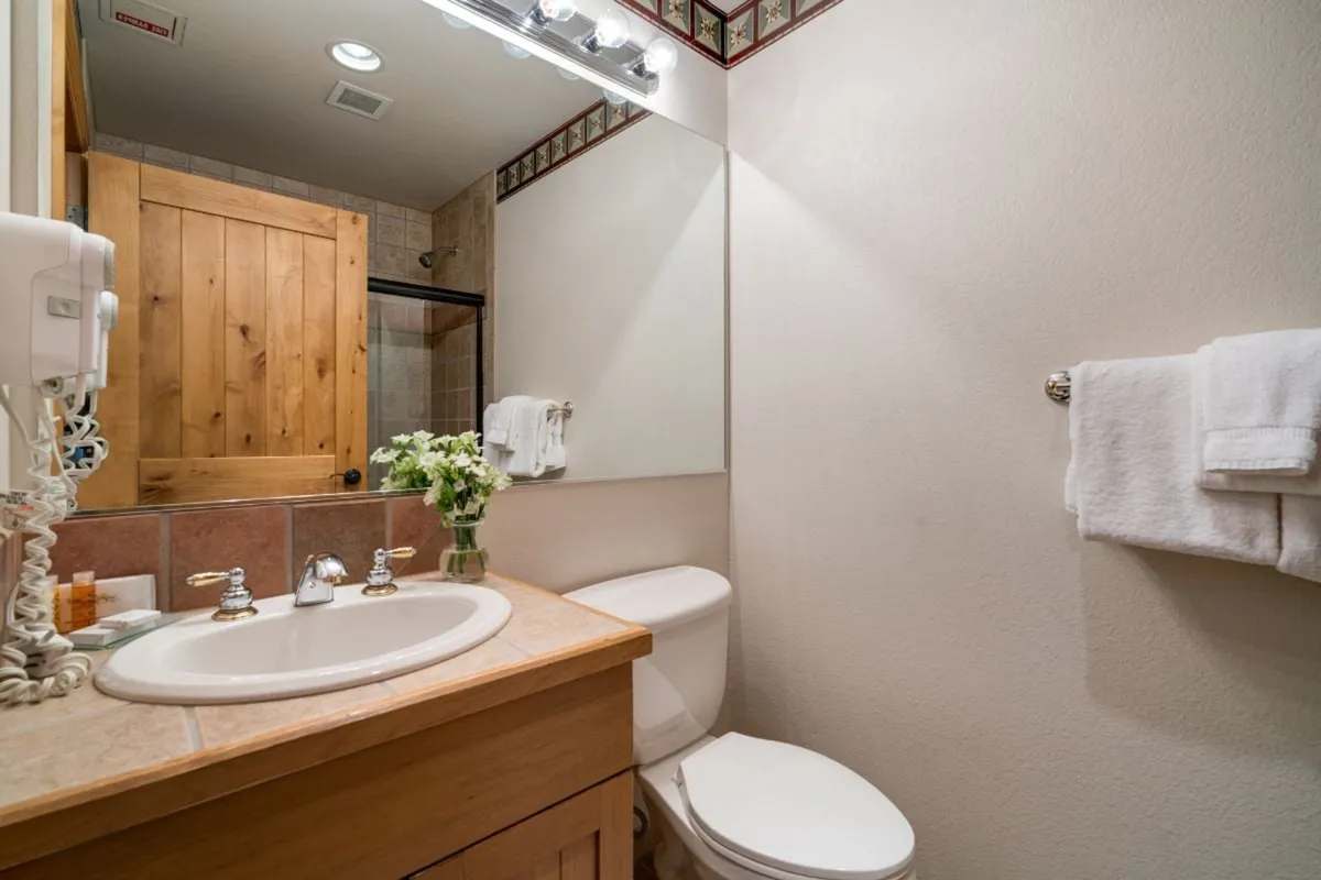 A serene corner of a bathroom displays a single sink with a stylish wooden cabinet, neatly arranged towels, and a delicate flower arrangement. The calming, uncluttered space creates an inviting environment for personal relaxation and self-care.