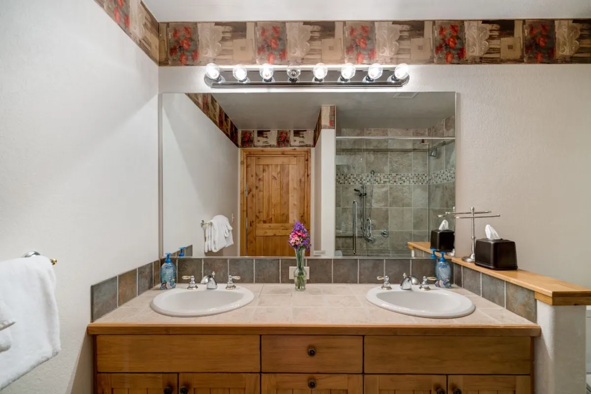 A modern ensuite bathroom with a double-sink vanity, warm wooden cabinetry, decorative wallpaper, a large mirror, and a glimpse of the walk-in shower. Fresh flowers and calm lighting create a spa-like, inviting atmosphere.