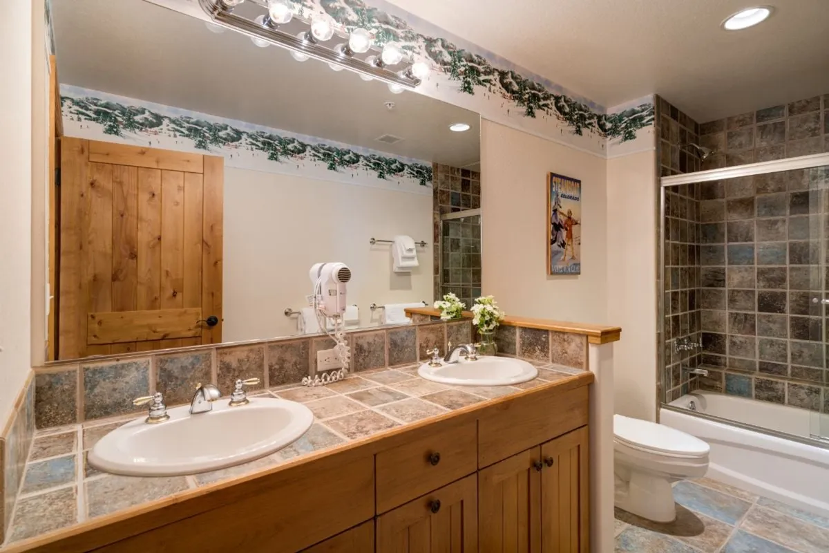 A spacious bathroom features a double sink with elegant wooden cabinetry and a decorative border, leading to a walk-in shower that evokes a spa-like ambiance. The neutral tones and thoughtful layout offer a sense of tranquility and privacy, ideal for relaxing after a day of adventure.
