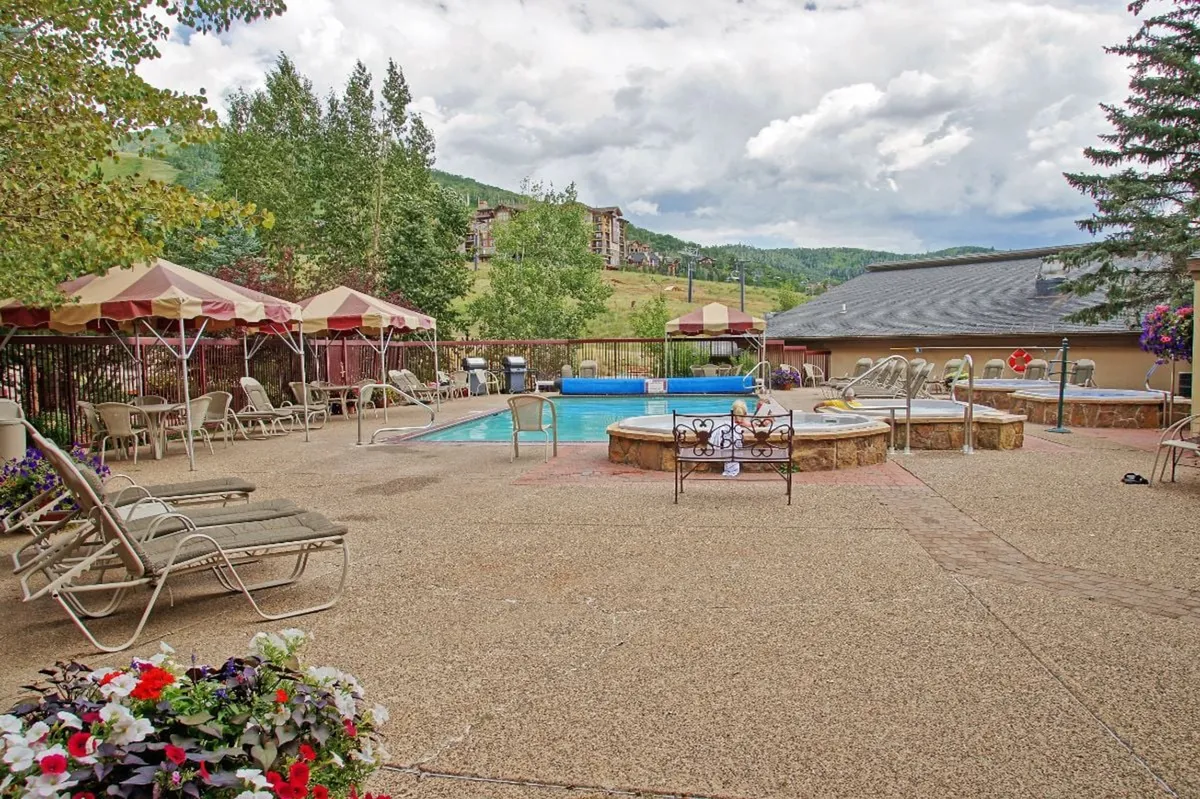 This image captures the inviting outdoor pool area, featuring a sparkling swimming pool, adjacent hot tub, lounge chairs, umbrellas, and lush flower beds. The scene is lively yet serene, hinting at both relaxation and social gatherings under the sun.