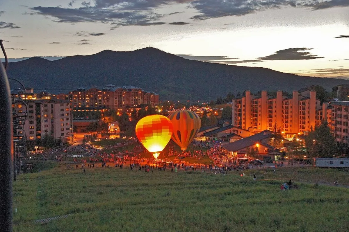 A vibrant outdoor scene showcasing hot air balloons ascending against a backdrop of mountains and illuminated buildings. There’s a lively crowd, suggesting a festive atmosphere with community gatherings and evening celebrations.