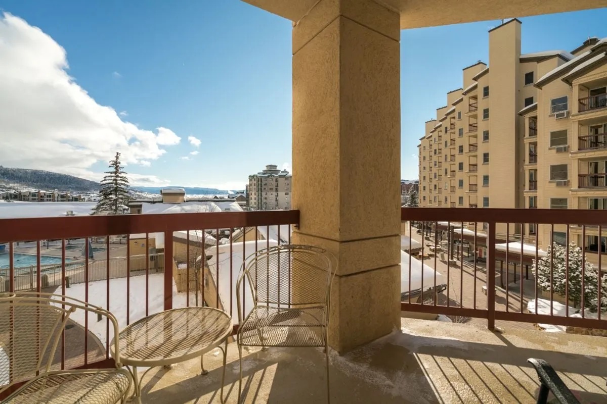 A serene, covered balcony with comfortable seating overlooks a snow-dusted landscape and distant mountains. The cozy yet airy space invites morning coffee or evening drinks while taking in scenic views of the base area year-round.