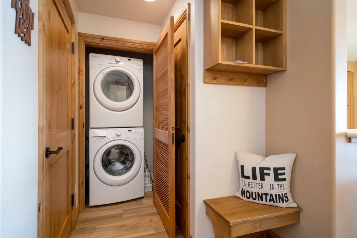 A practical laundry area featuring a stacked washer and dryer, a wooden bench with a soft cushion, and a convenient wooden shelf for organization. The space is both inviting and functional, offering the comforts of home.
