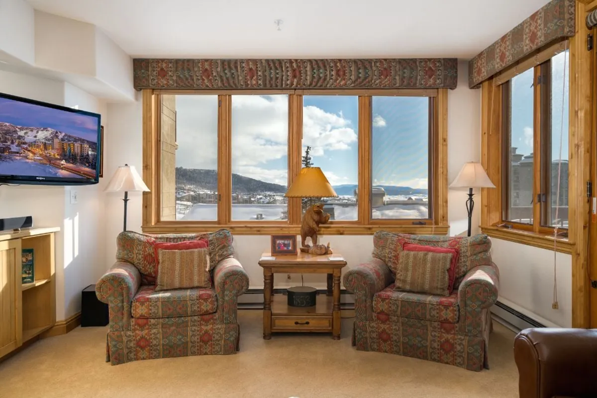 Warm, inviting sitting area with large windows, comfortable armchairs, a wooden coffee table, a decorative lamp, and a TV. The space is open and uncluttered, ideal for relaxing after outdoor activities.
